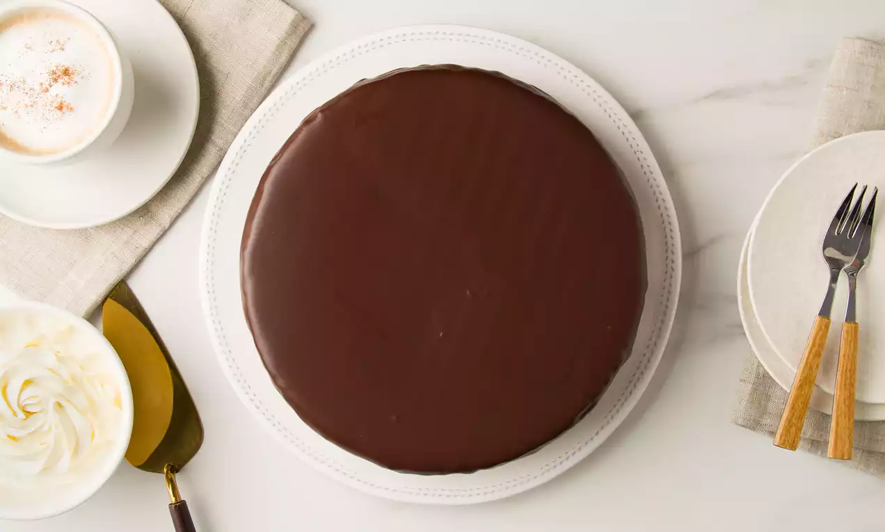 Tarta circular cubierta de brilloso glaseado de chocolate oscuro sobre plato blanco, acompañada de nata batida y café espolvoreado
