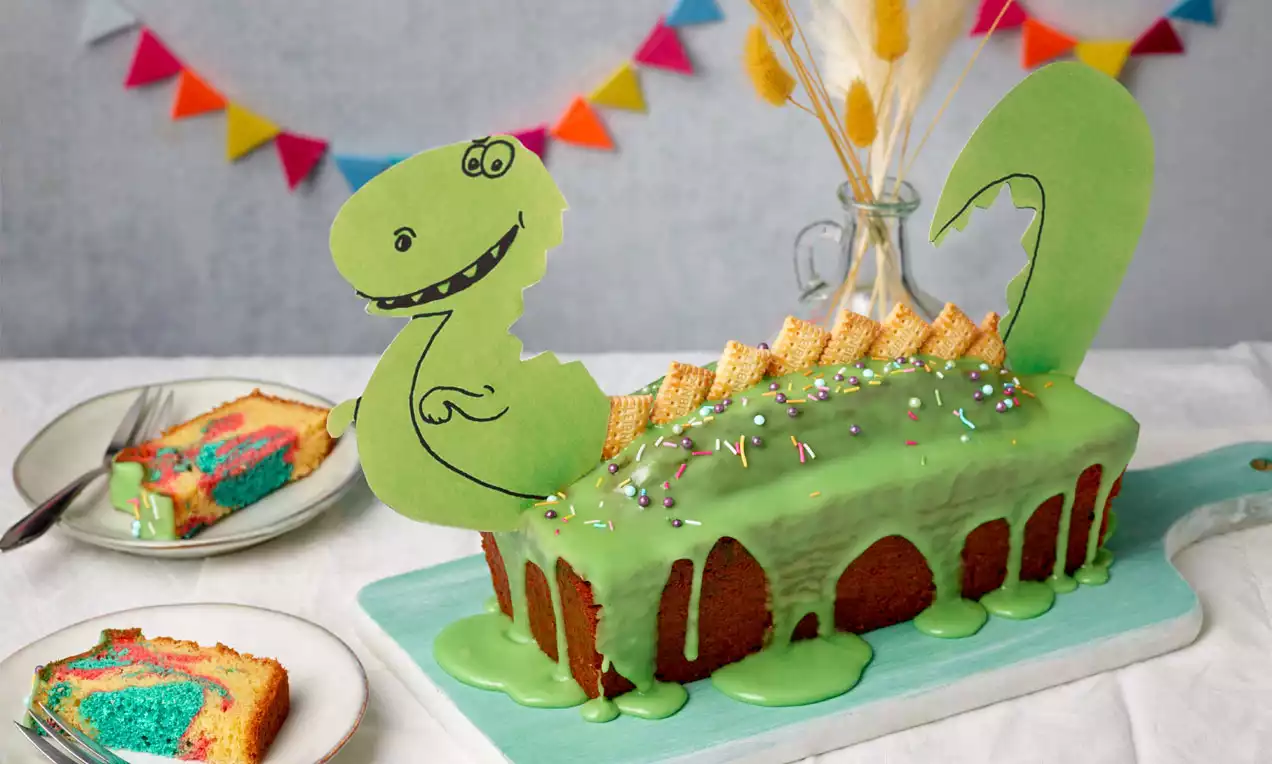 Dinokuchen mit grünem Zuckerguss und bunten Streuseln, verziert mit Waffelplatten und Papier-Deko, auf einem blauen Brett angerichtet