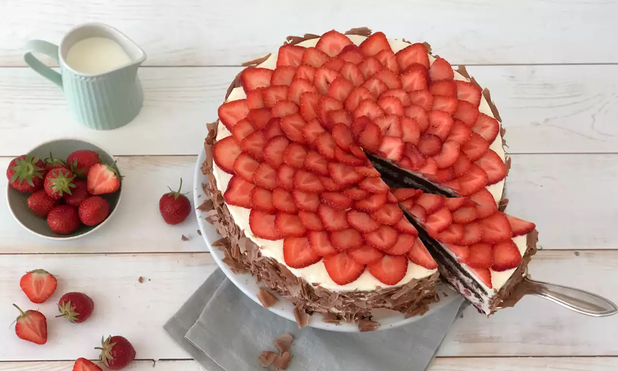 Schwarzwälder Erdbeertorte mit dichtem Erdbeerfächer auf weißer Sahne, Schokoraspeln am Rand, serviert auf Tortenteller mit grauer Stoffserviette