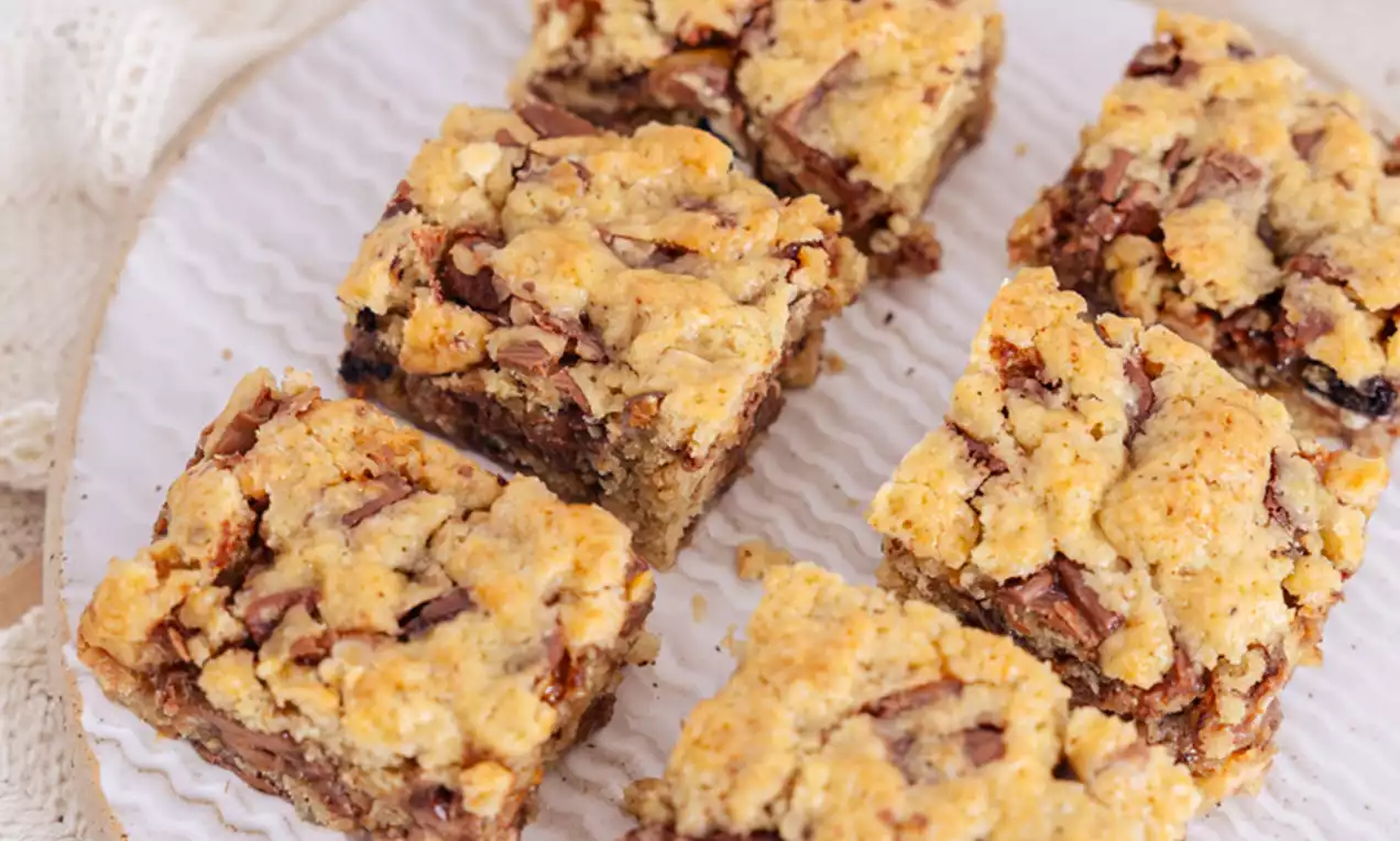 Zlate, kvadratne piškotne kocke z drobljivo zgornjo plastjo, prepredene s koščki čokolade na belem rebrastem krožniku