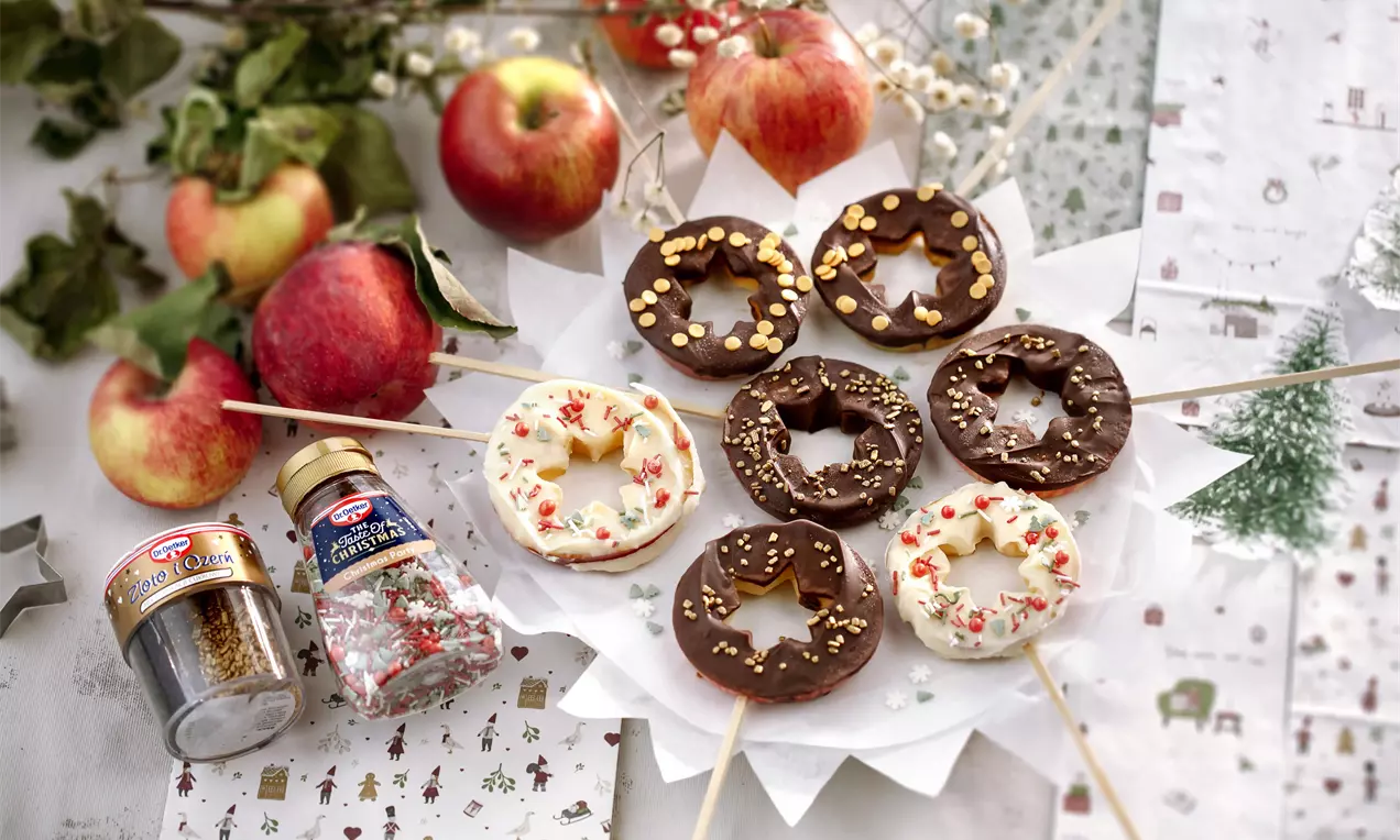 Apvalios obuolių riekelės su šokolado glaistu, puoštos pabarstukais, sudėtos ant baltos lėkštės, šalia raudonų obuolių ir dekoracijų