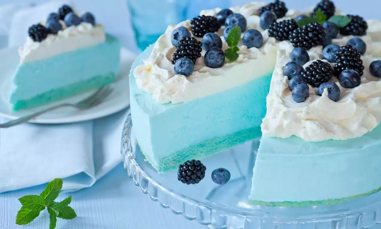 Błękitny sernik jogurtowy na przezroczystej paterze, z puszystym kremem waniliowym, udekorowany jagodami, jeżynami i listkami mięty