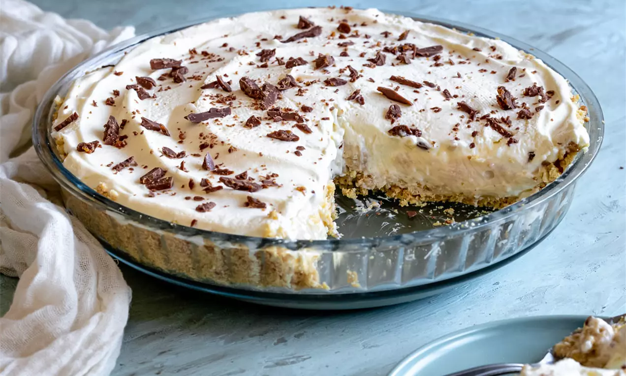 Бананов пай с кремообразна бяла плънка в стъклена тава върху бисквитен блат, поръсен с тъмнокафяви шоколадови стърготини