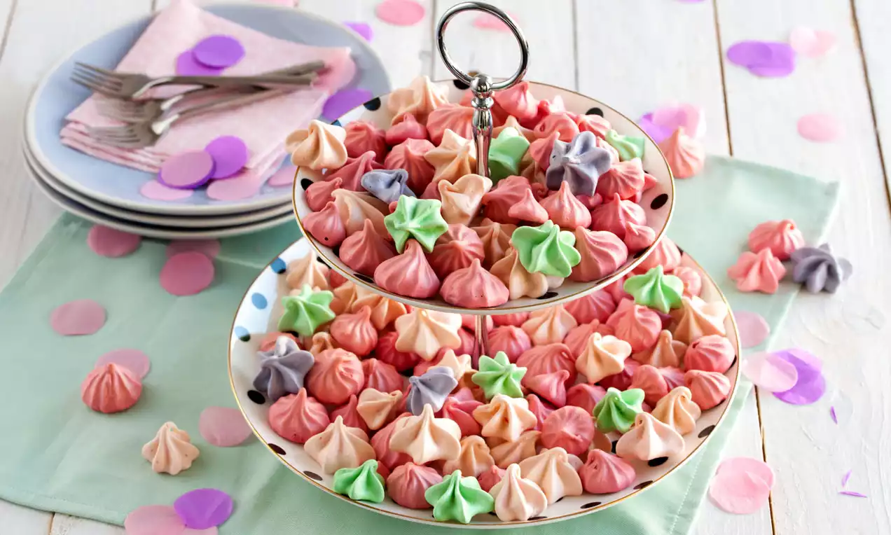 Bunte Meringue-Küsse in Rosa, Grün, Lila und Weiß mit weicher Textur auf einer zweistöckigen Etagere, umgeben von Dekokonfetti
