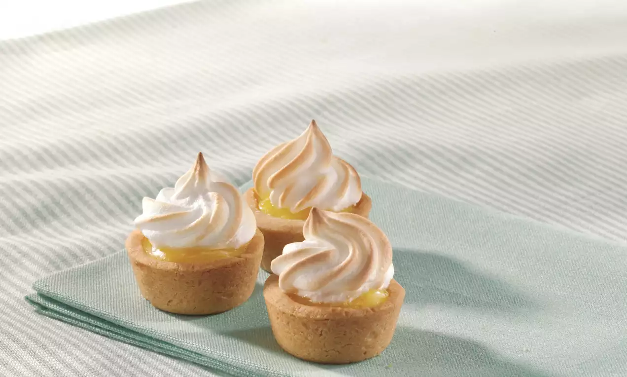 Mini lemon meringue pies with golden pastry crusts, swirled white and caramel meringue tops, arranged on a pale green cloth