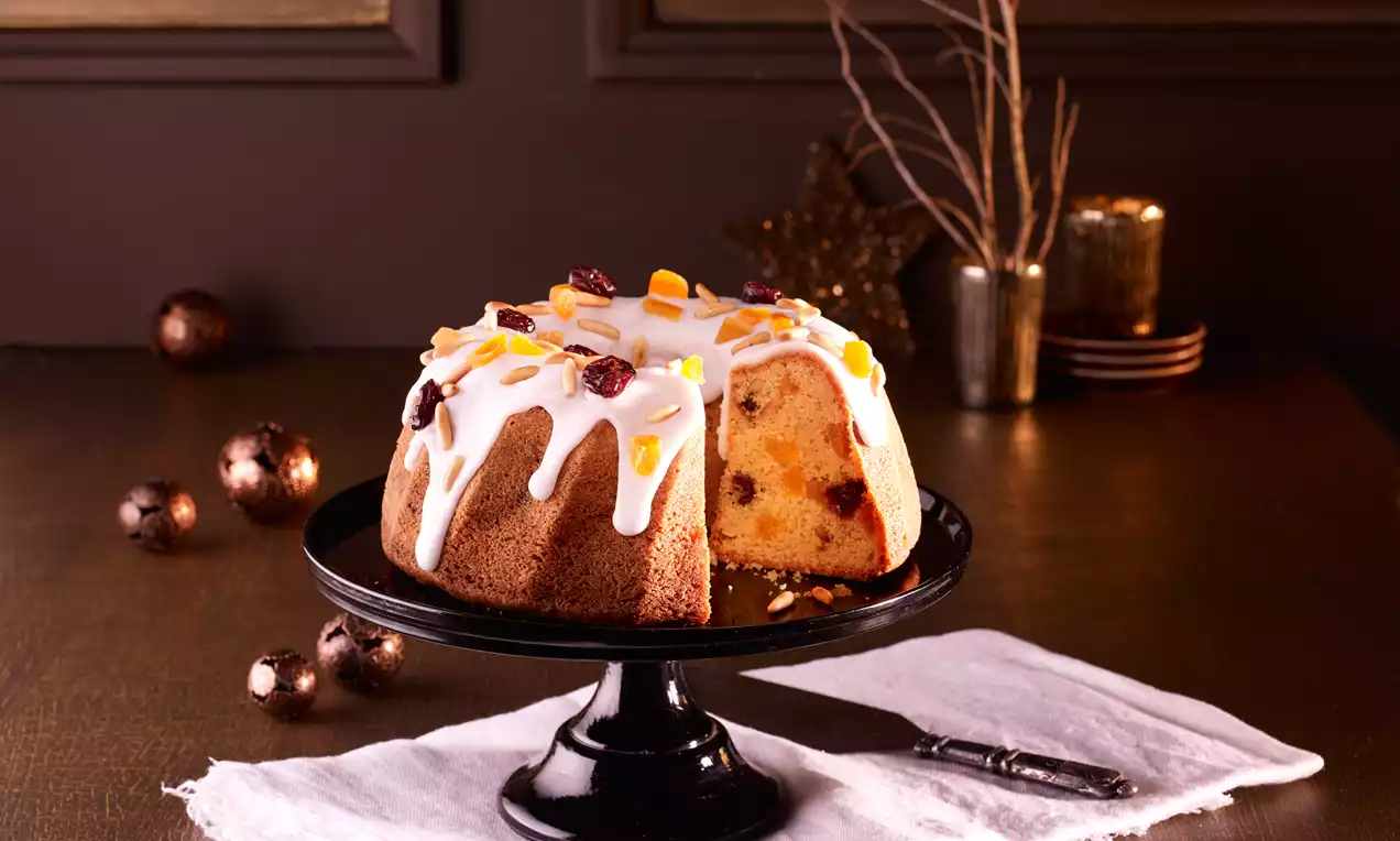 Torta a ciambella dorata con glassa bianca lucida colata, decorata con scorze candite e frutta secca su un’alzata nera e tovaglietta bianca