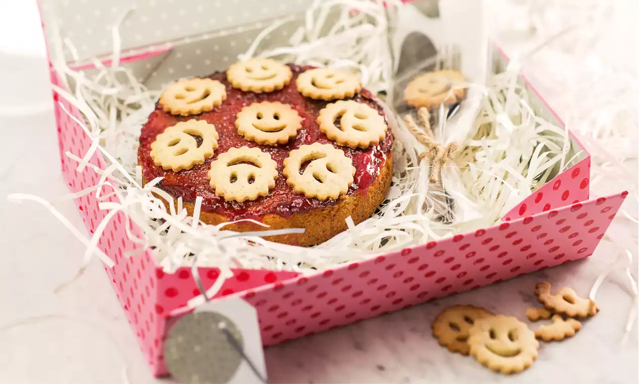 Runde, weiße Mini-Linzer-Torte mit roter Konfitüre, verziert mit lächelnden Keksdeckeln, gebettet in weiße Deko-Fasern in gepunkteter Box
