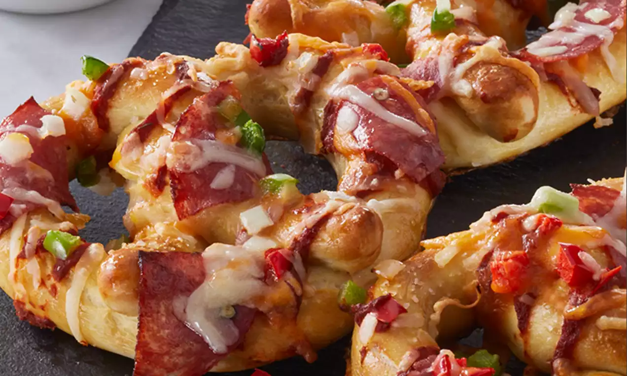 Golden brown pretzels covered in melted cheese, sliced pepperoni, and colorful diced peppers on a slate with a small bowl of red marinara