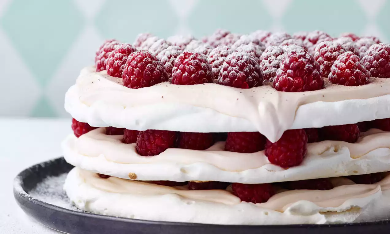 Hvit marengskake lagdelt med rosa bringebærkrem og friske røde bringebær, drysset med melis, på mørk tallerken mot diamantmønstret bakgrunn