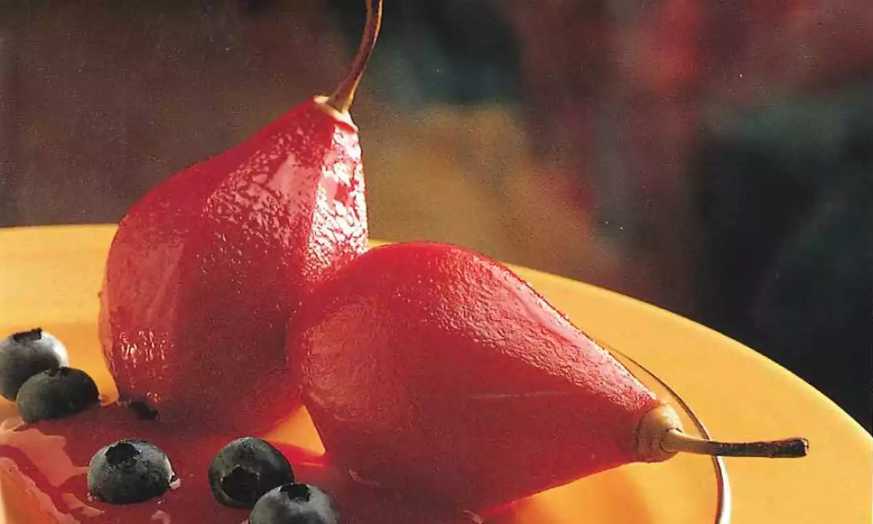 Twee glanzende, dieprode, gepocheerde peren met steeltjes in een glanzende rode saus, gegarneerd met blauwe bessen op een geel bord