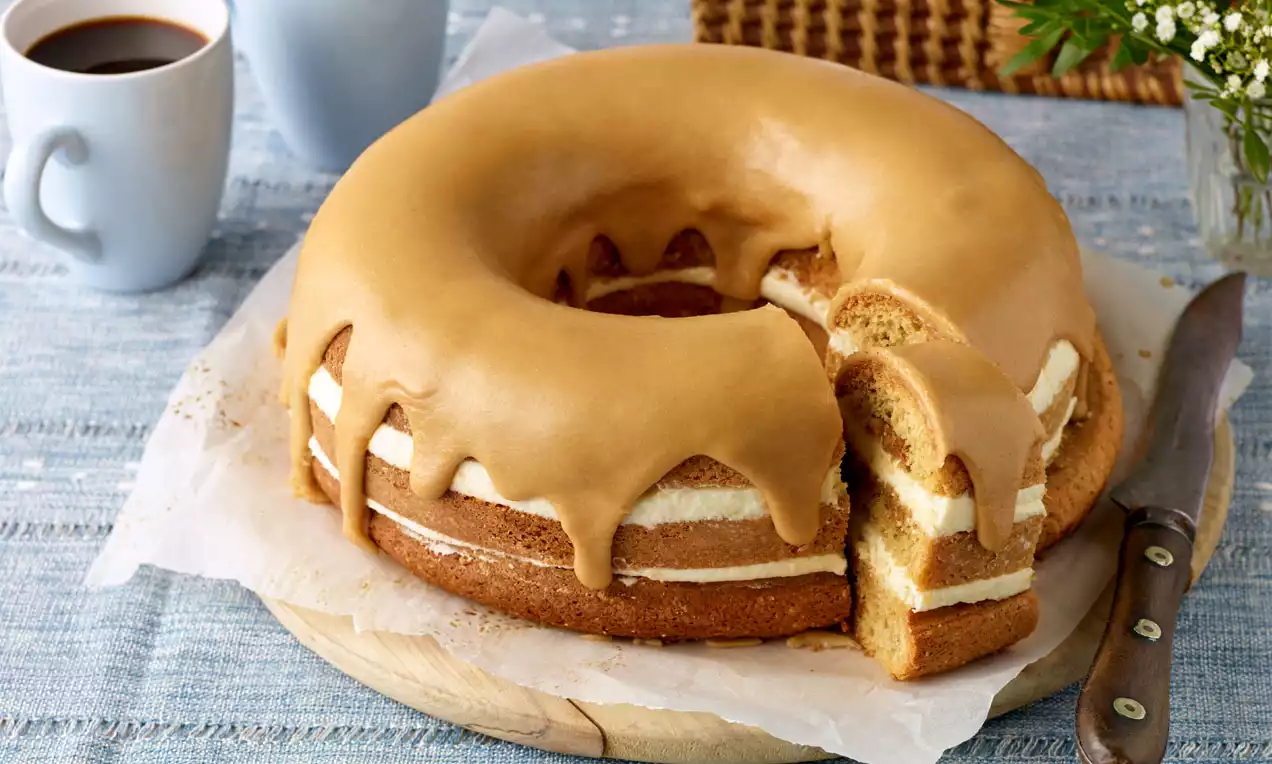 Vaaleanruskea kuorrute valuu pehmeän pyöreän kakun päältä, jonka välistä näkyy kermaista täytettä, aseteltuna vaalealle puualustalle