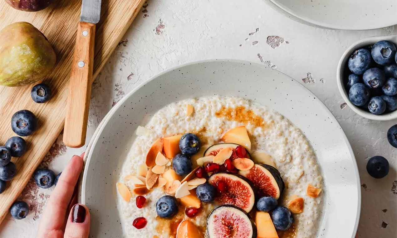 Picture - Creamy white quinoa porridge topped with colorful fresh figs, blueberries, papaya, pomegranate seeds, and almond flakes in a shallow bowl