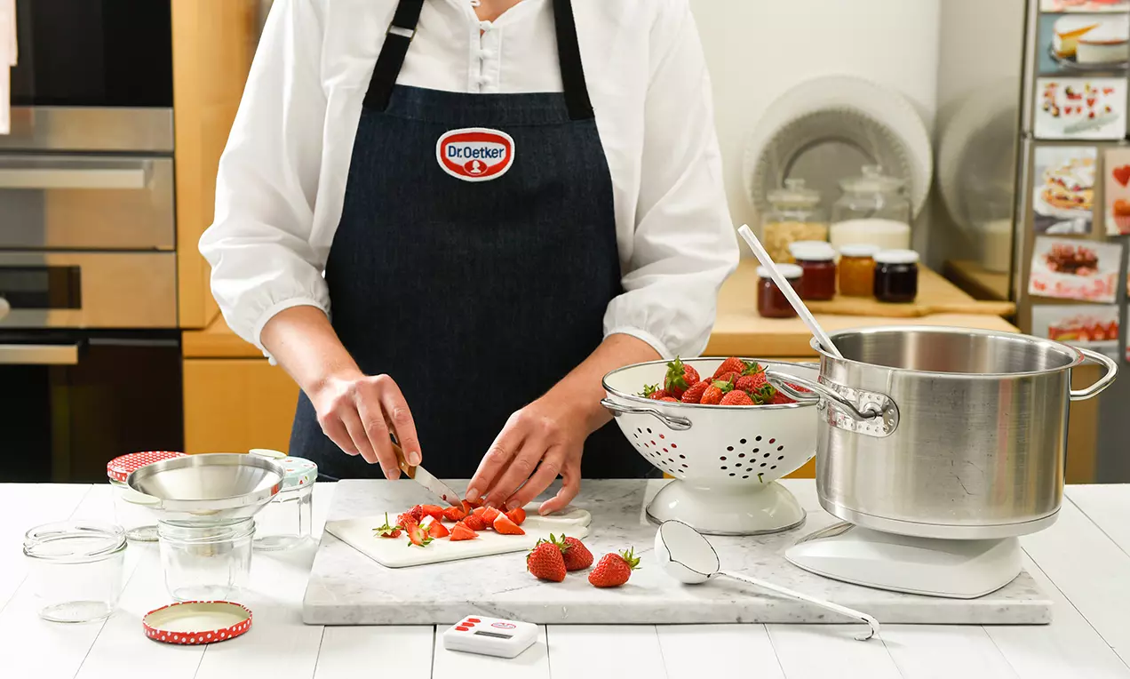 Na bielej doske sa krájajú jasne červené jahody, vedľa kovového sitka plného jahôd, veľkého hrnca a pripravených sklenených pohárov