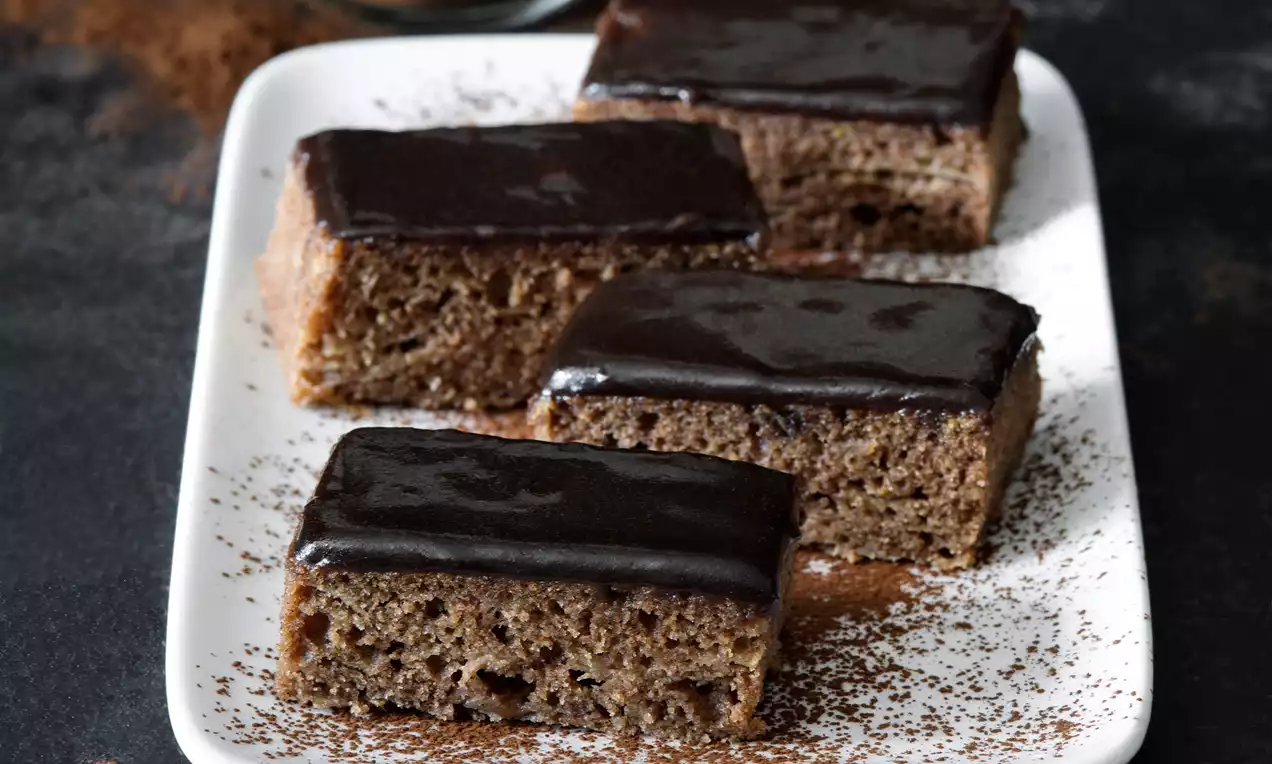 Čtvercové kousky karobového jablečného perníku s tmavou lesklou polevou na bílém talíři sypaném jemným kakaovým práškem