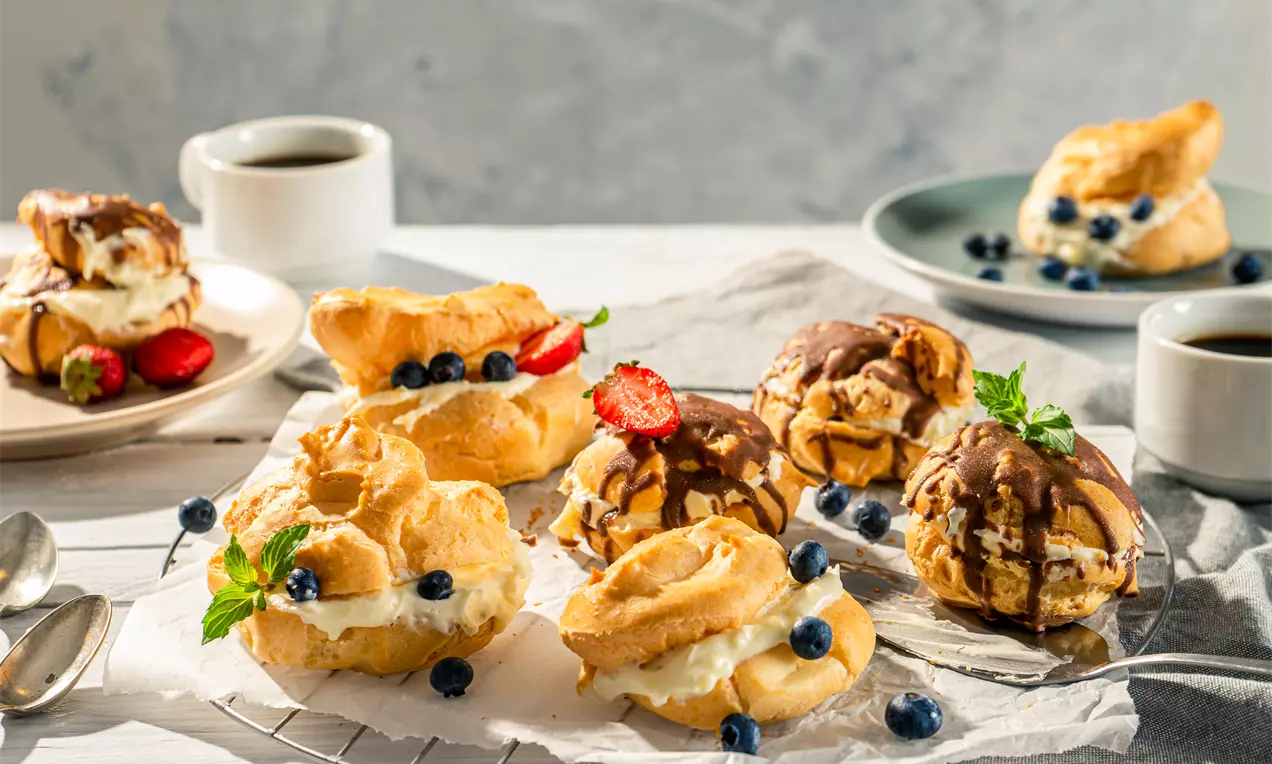 Еклери със златиста и леко напукана повърхност, пълни с ванилов крем, гарнирани с пресни плодове и шоколад, подредени върху бяла хартия