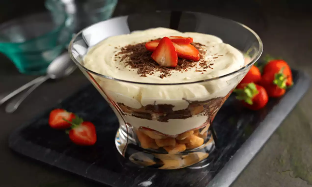 Layered trifle in a glass bowl with mascarpone cream, sponge fingers, chocolate shavings, and halved strawberries on top