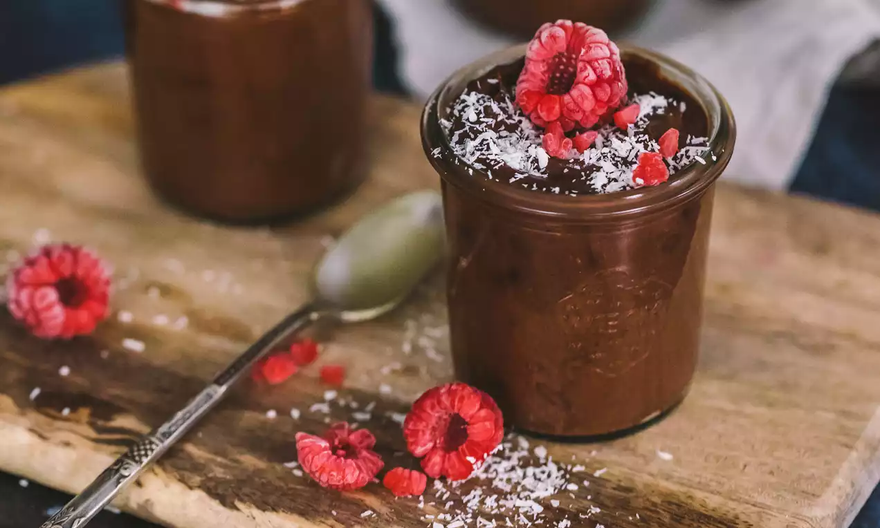 Mousse vegana de chocolate oscuro y cremoso servido en un vaso, decorado con frambuesas frescas y coco rallado sobre tabla de madera