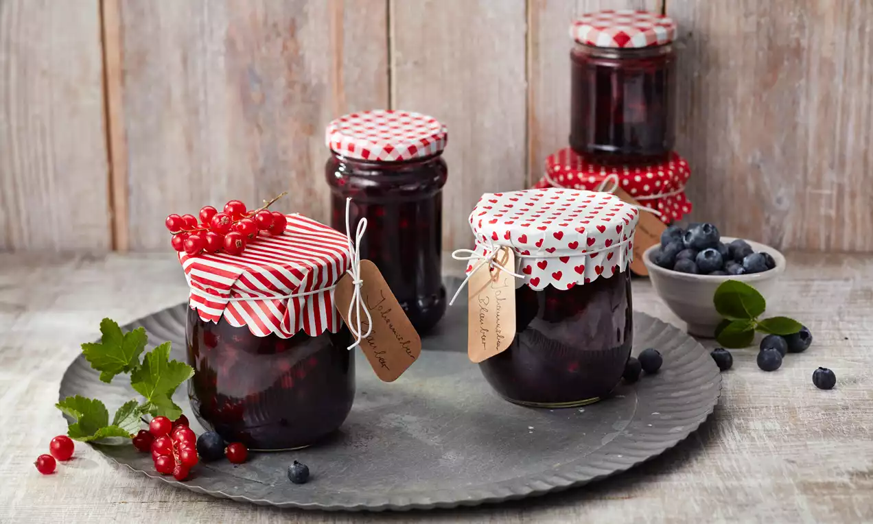 Gläser mit dunkelroter Konfitüre, verziert mit rot gemusterten Deckeln und Anhängern, arrangiert auf einem Zinnteller mit Beeren und Blättern