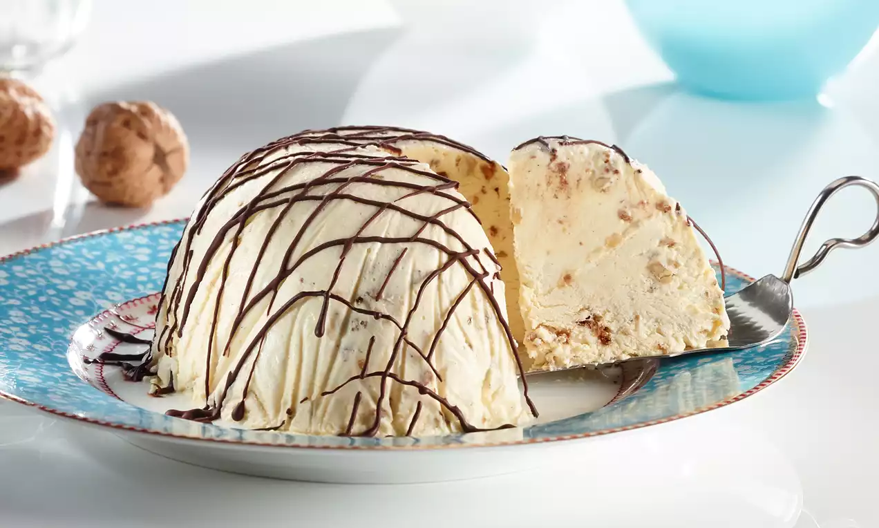 Dôme glacé crème ivoire parsemé de morceaux de noix avec un nappage de filets de chocolat noir sur une assiette ornée de motifs bleus