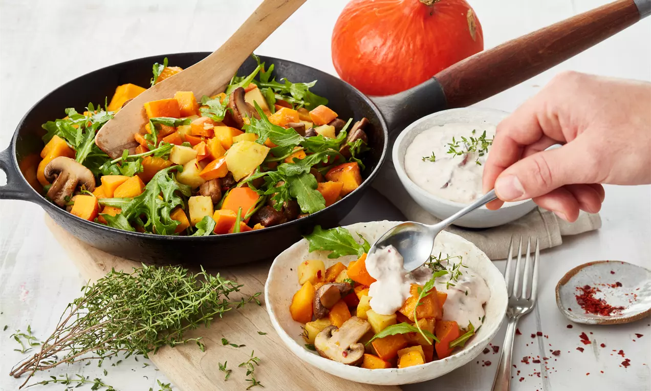 Bunte Kürbis-Pfanne mit orangefarbenen Kürbisstücken, Zucchini, Pilzen, Rucola und frischem Thymian in schwarzer Pfanne, angerichtet mit Creme-Dip