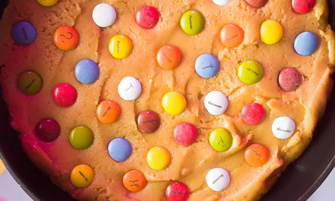 Galleta dorada y gruesa con superficie uniforme decorada con Lacasitos de colores vivos dispuestos de forma irregular dentro de una sartén negra