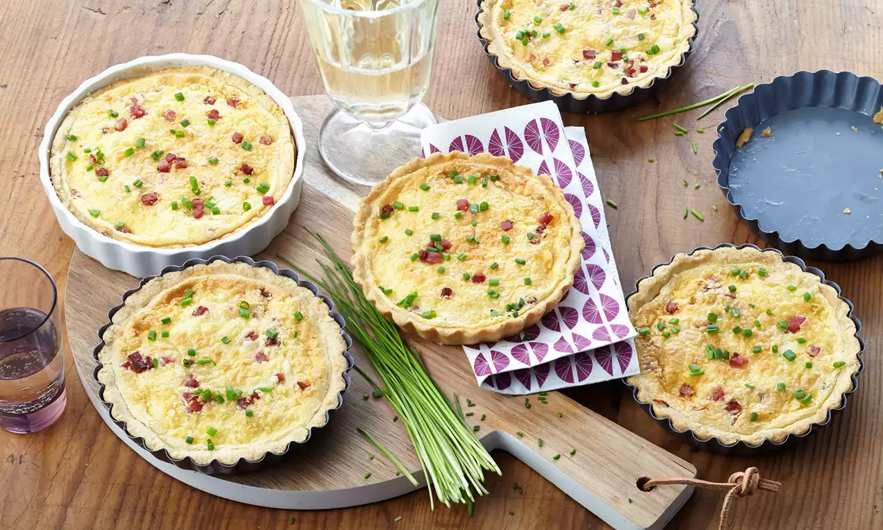 Goldgelbe Mini-Quiches Lorraine in gewellten Förmchen mit knusprigem Rand, bestreut mit grünem Schnittlauch auf einem Holzbrett angerichtet