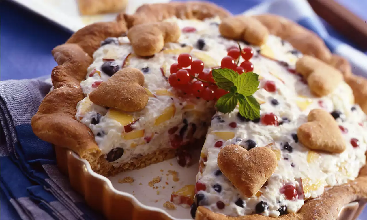 Runder Kuchen mit goldbraunem Rand, Fruchtreisfüllung mit bunten Fruchtstücken, Herzkeksen, Minzblatt und roten Johannisbeeren garniert