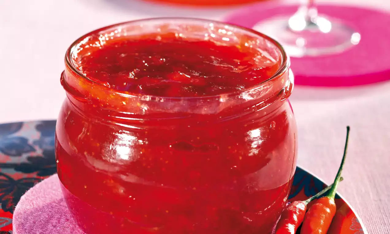 Erdbeerrote Konfitüre mit glänzender Oberfläche in einem gläsernen Einmachglas, daneben zwei rote Chilischoten auf einem bunten Teller