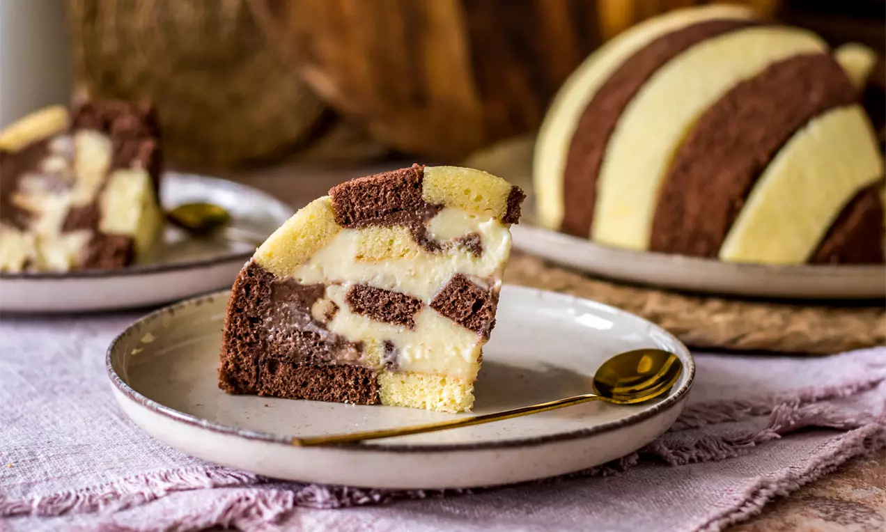 Торта с редуващи се райета от светложълт и какаов блат върху кремообразен пълнеж в напречен разрез, сервирана върху светла чиния с лъжичка