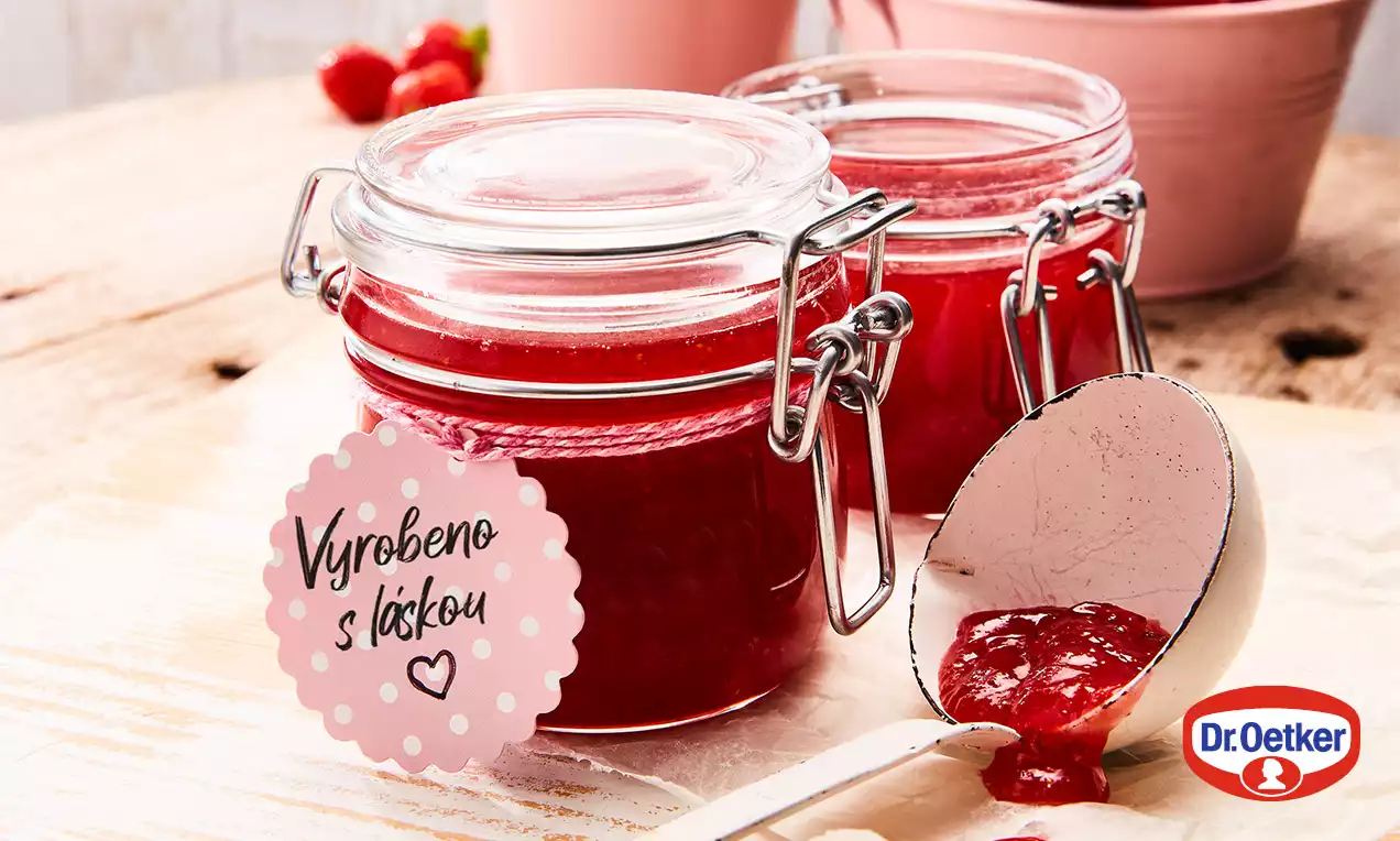 Sklenice plné sytě červeného džemu s hladkou texturou, uzavřené víčkem, zdobené růžovým štítkem, vedle bílé lžíce s porcí džemu