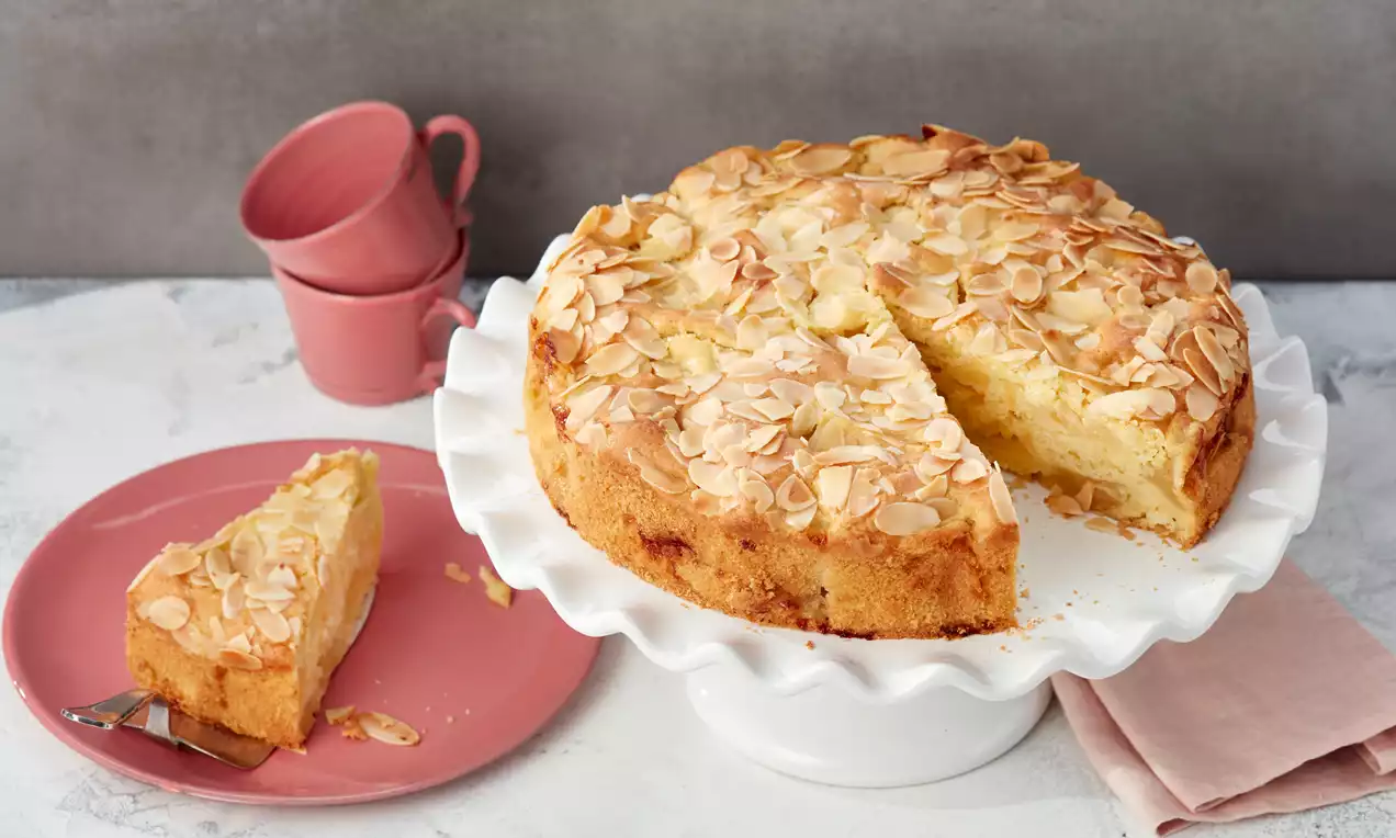 Veganer Apfelkuchen mit goldbrauner Kruste, roten Apfelscheiben, knusprigen Mandelblättchen und Puderzucker auf hellem Teller serviert