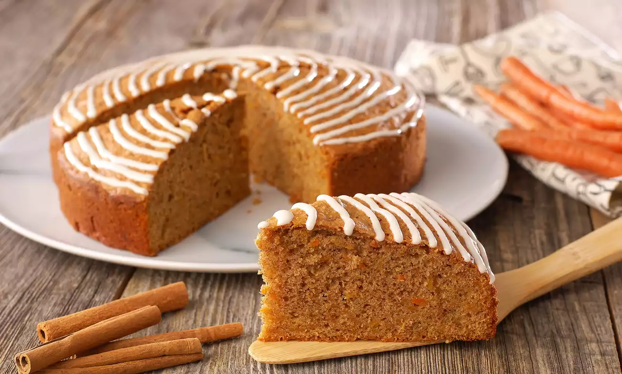 Tam buğday unlu havuçlu kek, tarçın çubukları ve taze havuçlarla süslenmiş, üstü beyaz glazür şeritleriyle desenlenmiş rustik sunum