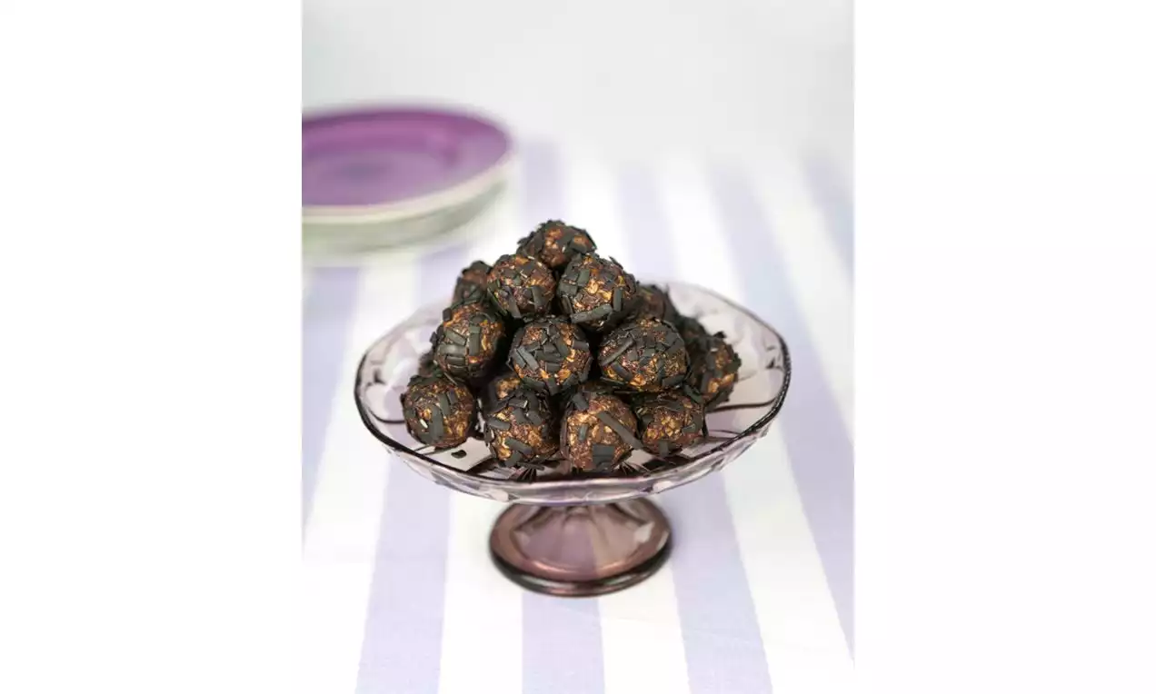 Lakritsbollar med mörk yta och krispiga bitar, staplade i pyramidform på ett lila glasfat, stående på en ljusrandig duk