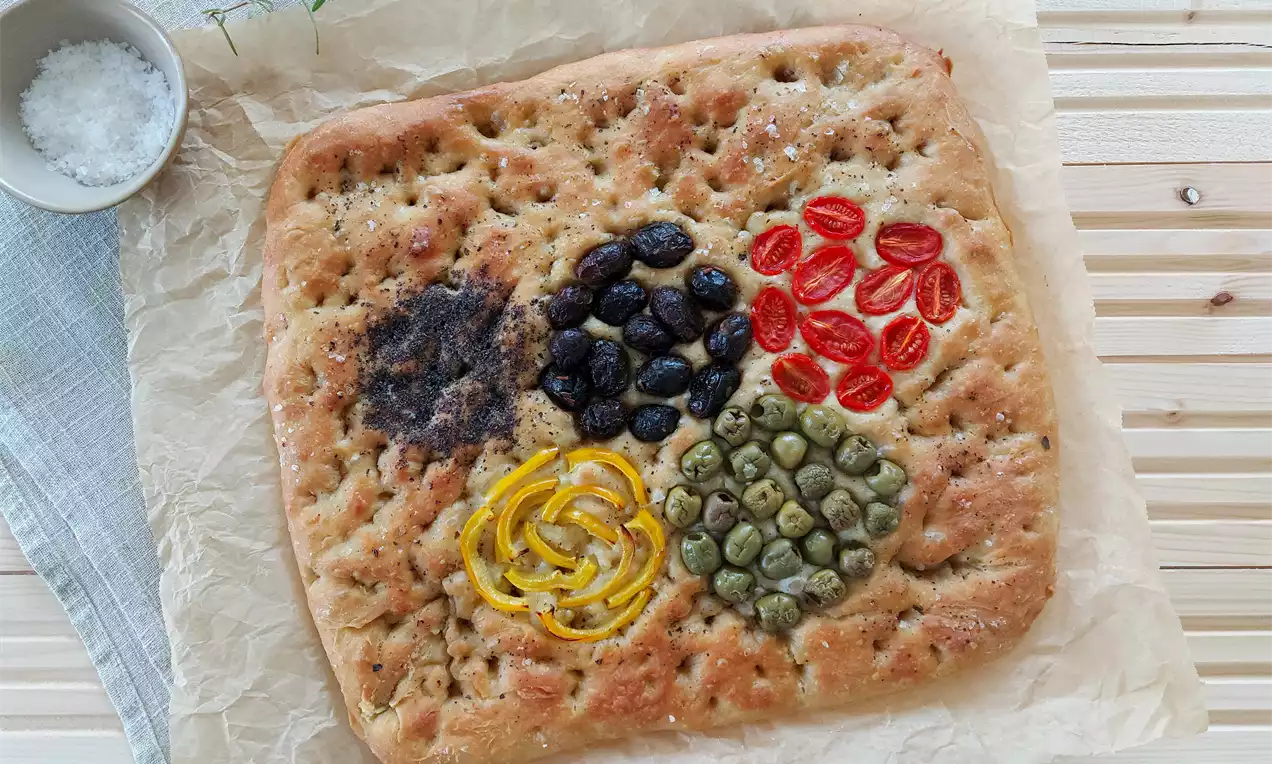 Zlatohnědá plochá focaccia s měkkou texturou, zdobená olivami, rajčaty, citrusovou kůrou, kapary a kořením na pečicím papíru