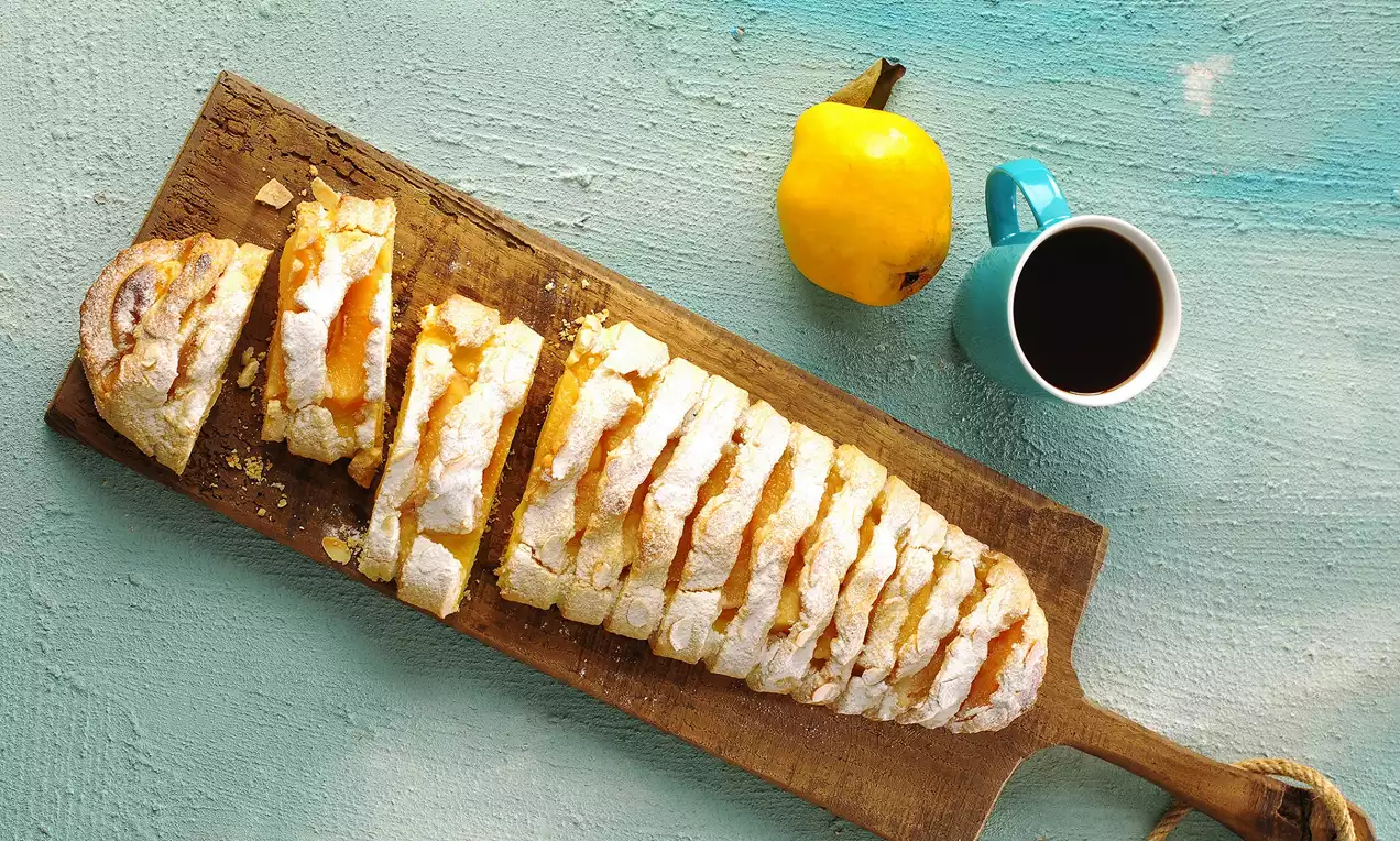 Ahşap bir sunum tahtası üzerinde dilimlenmiş, üzeri pudra şekeri serpilmiş altın sarısı ayvalı hamur tatlısı, yanında ayva ve bir fincan kahve