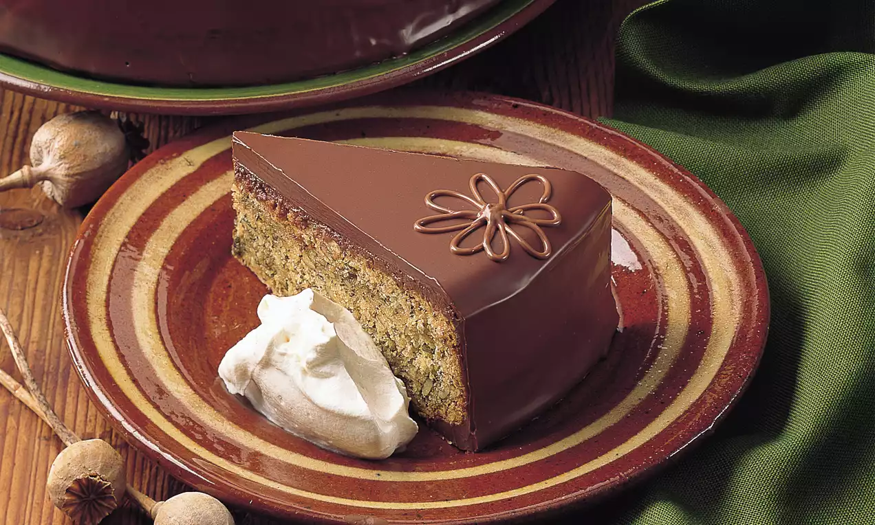 Dreieckiges Stück Kürbis-Mohn-Torte mit dunkler Schokoglasur und Schokoblume auf braunem Teller, daneben Klecks weißer Sahne