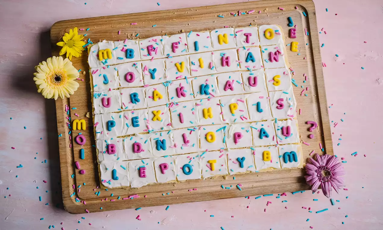 Neliöiksi leikatut vaaleat leivokset aseteltu puiselle alustalle, valkoisella kuorrutteella ja värikkäillä kirjainkoristeilla, vieressä kukkia