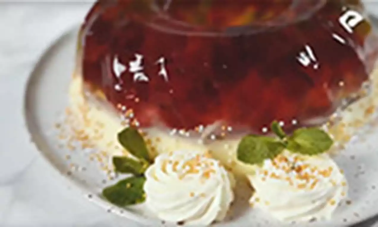 Gaiši baltā šķīvī uz zeltainu drupaču fona sārta želeja caurspīdīgā formā ar spirālveidīgām putukrējuma rozēm un svaigas piparmētras lapiņām