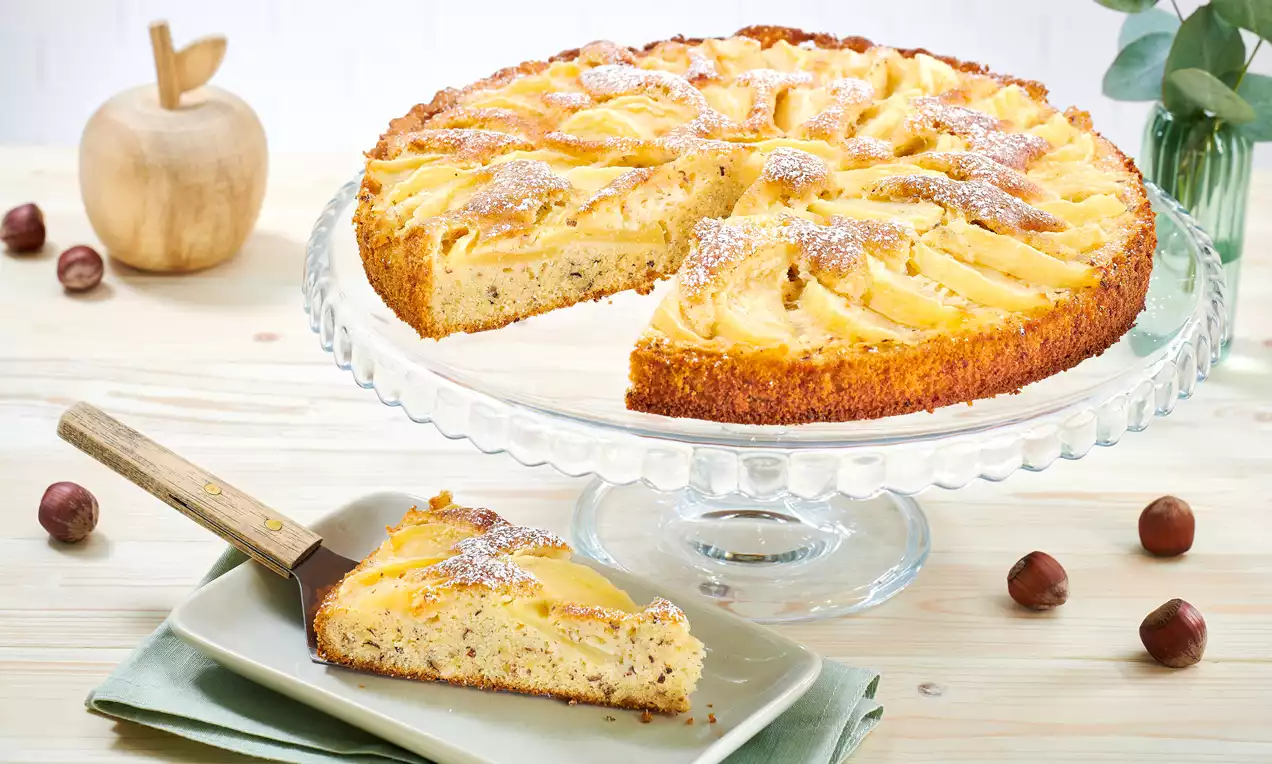 Apfel-Nuss-Tarte mit goldgelber Oberfläche und geschnittenen Apfelspalten auf Glasplatte, daneben ein Stück auf weißem Teller mit Gabel