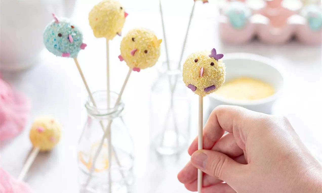 Малки кръгли кейк попс в пастелно жълто и синьо, с текстура от захарни поръски и очички, поставени на бели клечки в стъклена бутилка