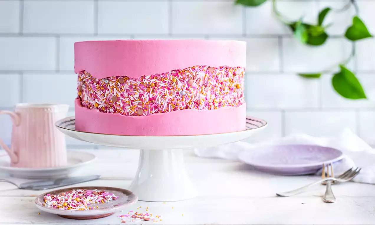 Roze Fault Line Cake met kleurrijke strooisels in een golvende band, op een wit taartplateau met zachtroze en paarse serviesstukken