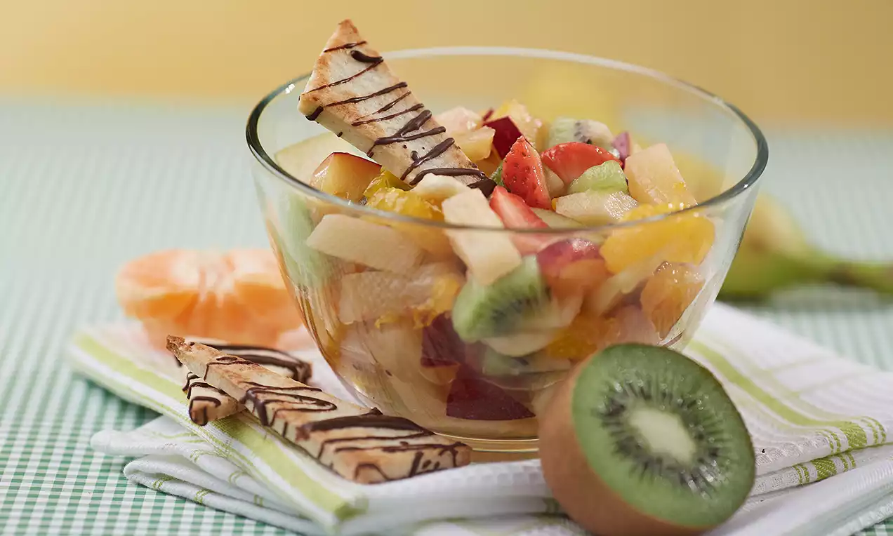 Bunter Fruchtsalat aus frischen Kiwi-, Erdbeer-, Apfel- und Orangenstücken in einer Glasschale, dekoriert mit Schoko-Gebäcksticks