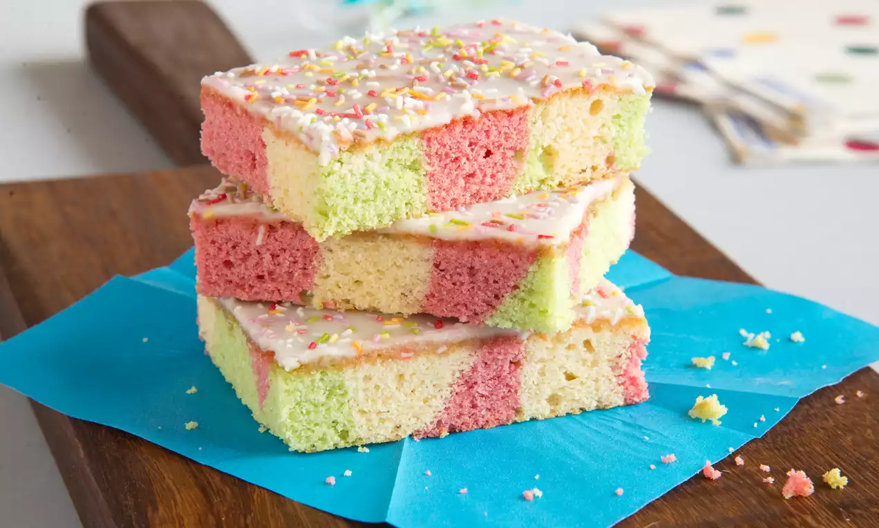 Papagajski kolač s kvadratnim pastelno ružičastim, žutim i zelenim slojevima, prekriven glazurom i šarenim mrvicama na plavom papiru