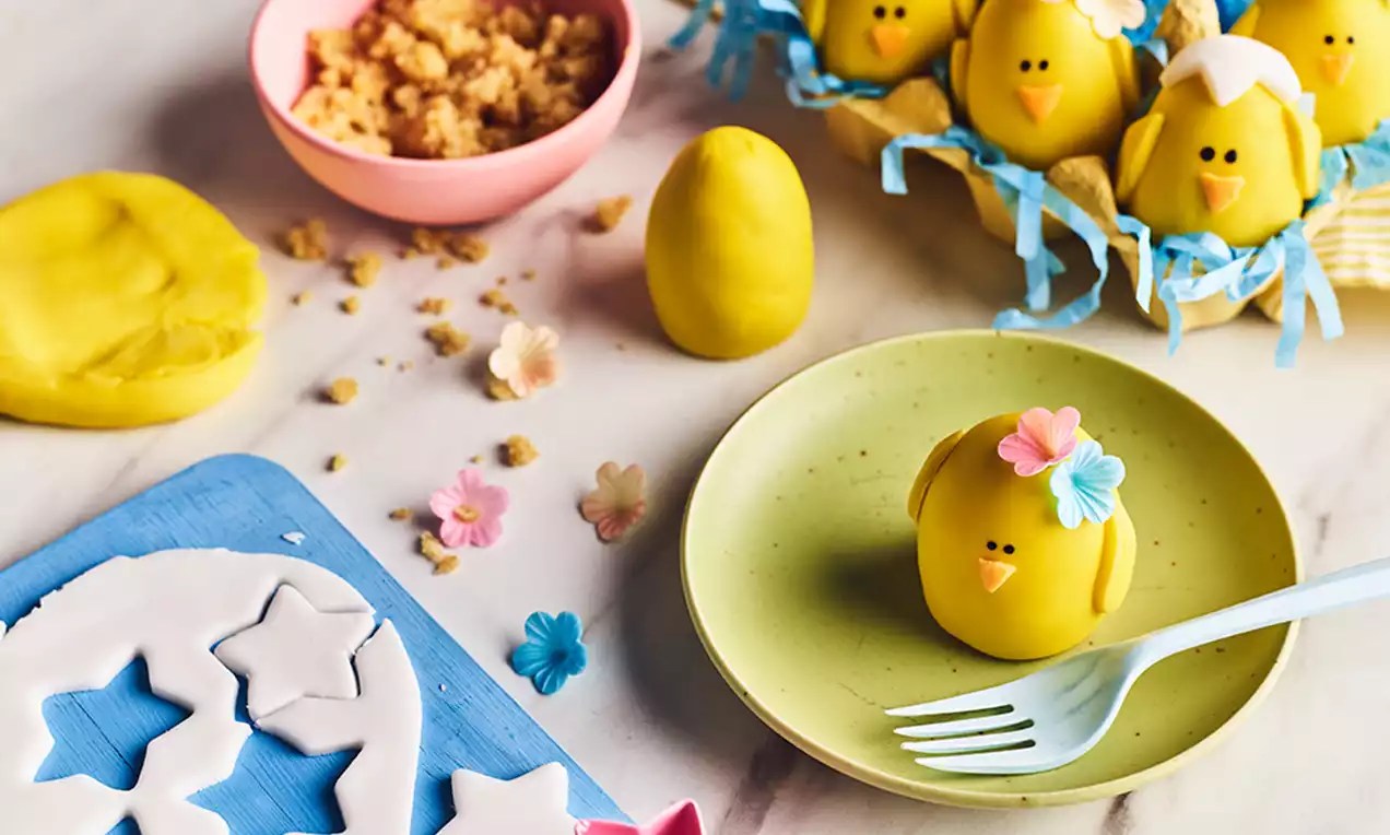 Gula marsipankycklingar med vita detaljer och blommor, formade som ägg på ett blått fat med pappersstrimlor och marsipanpyssel