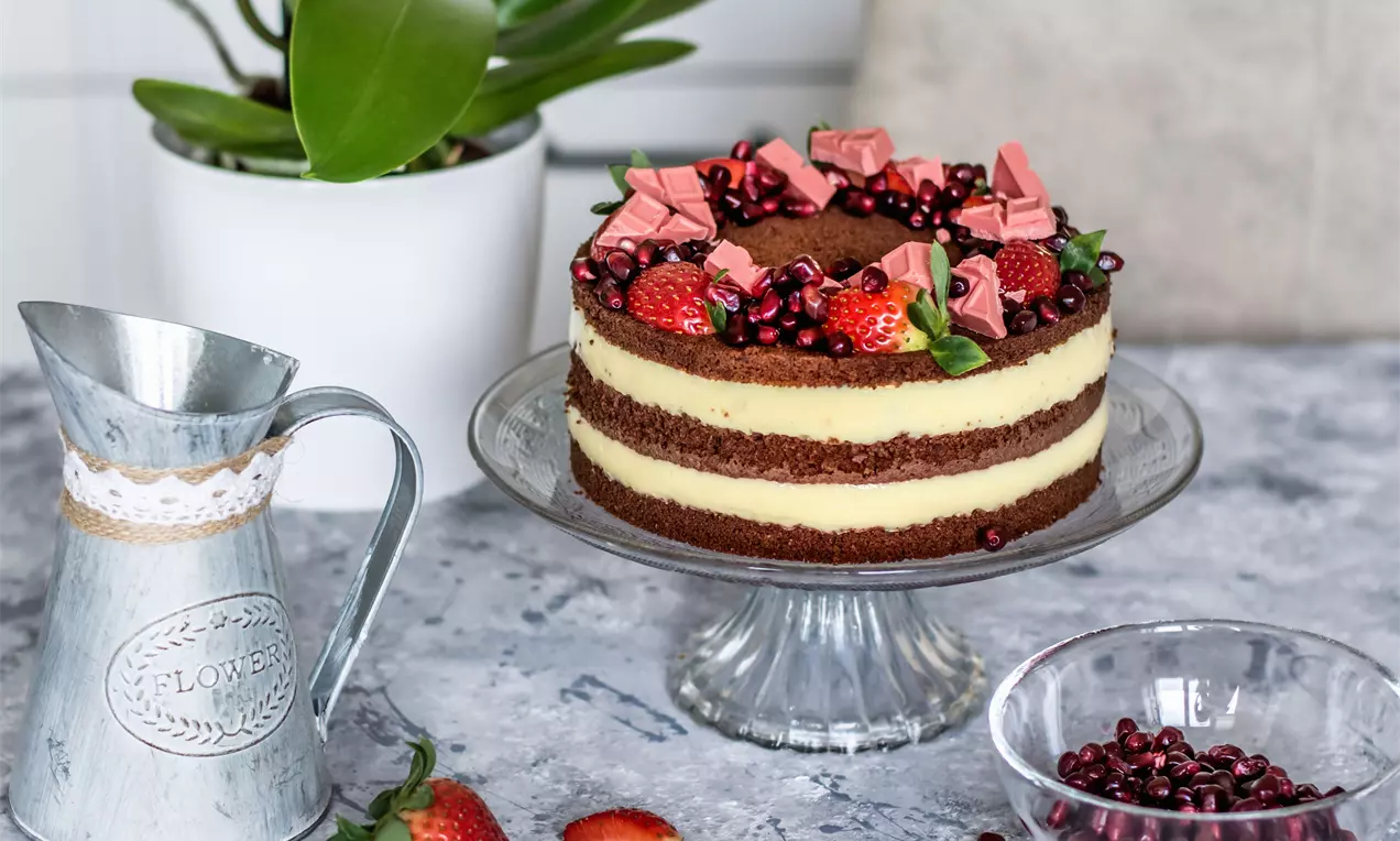 Бисквитена торта с шоколадови пластове и светъл ванилов крем, украсена с ягоди, нар, мента и розови шоколадови парчета върху стъклена стойка