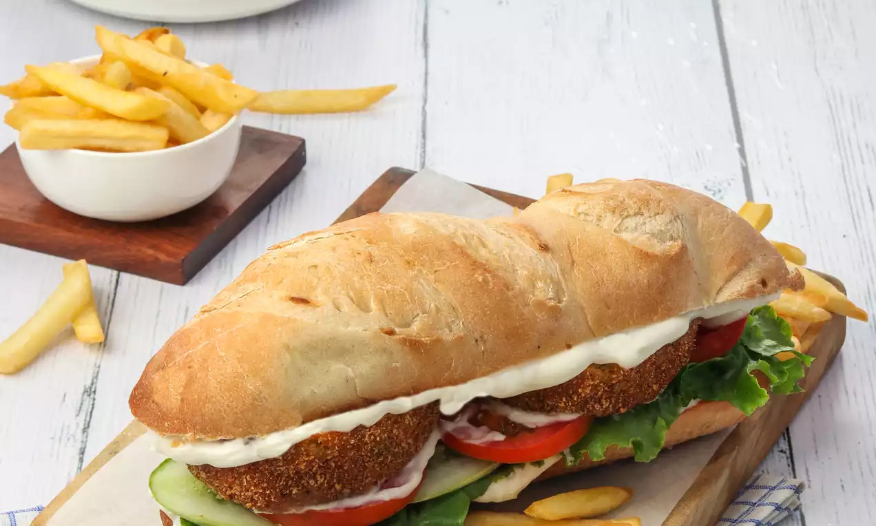 Golden brown baguette filled with crispy breaded potato patty, lettuce, tomato slices, and sauce, served on a wooden board with fries