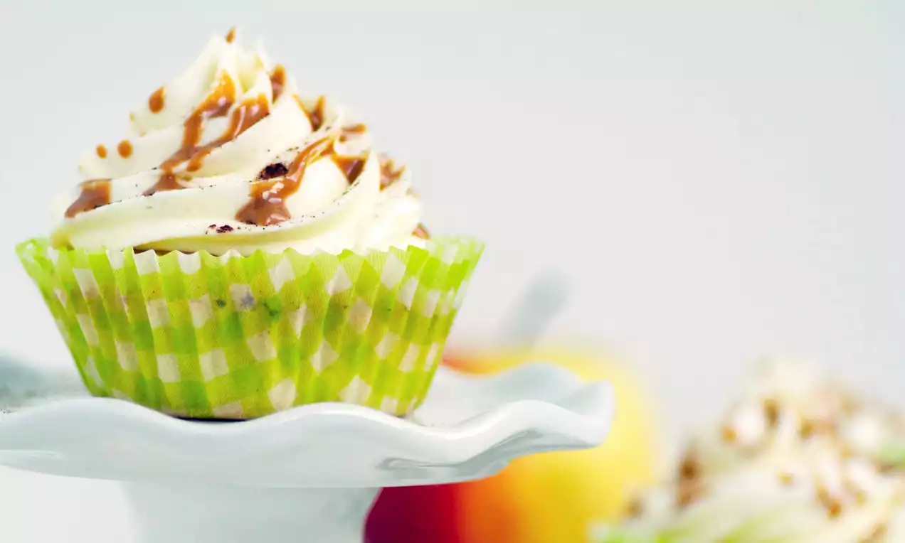 Winterse cupcakes met witte slagroomtoef en karamelsaus in groene geruite papieren vormpjes op een wit bord met appels op de achtergrond