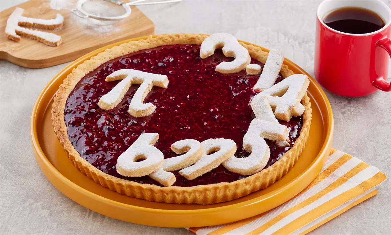 Altın rengi tabanlı tart, parlak kırmızı meyve dolgusu üzerinde pudra şekerli pi sayısı şekilleriyle sarı bir tabakta sunulmuş