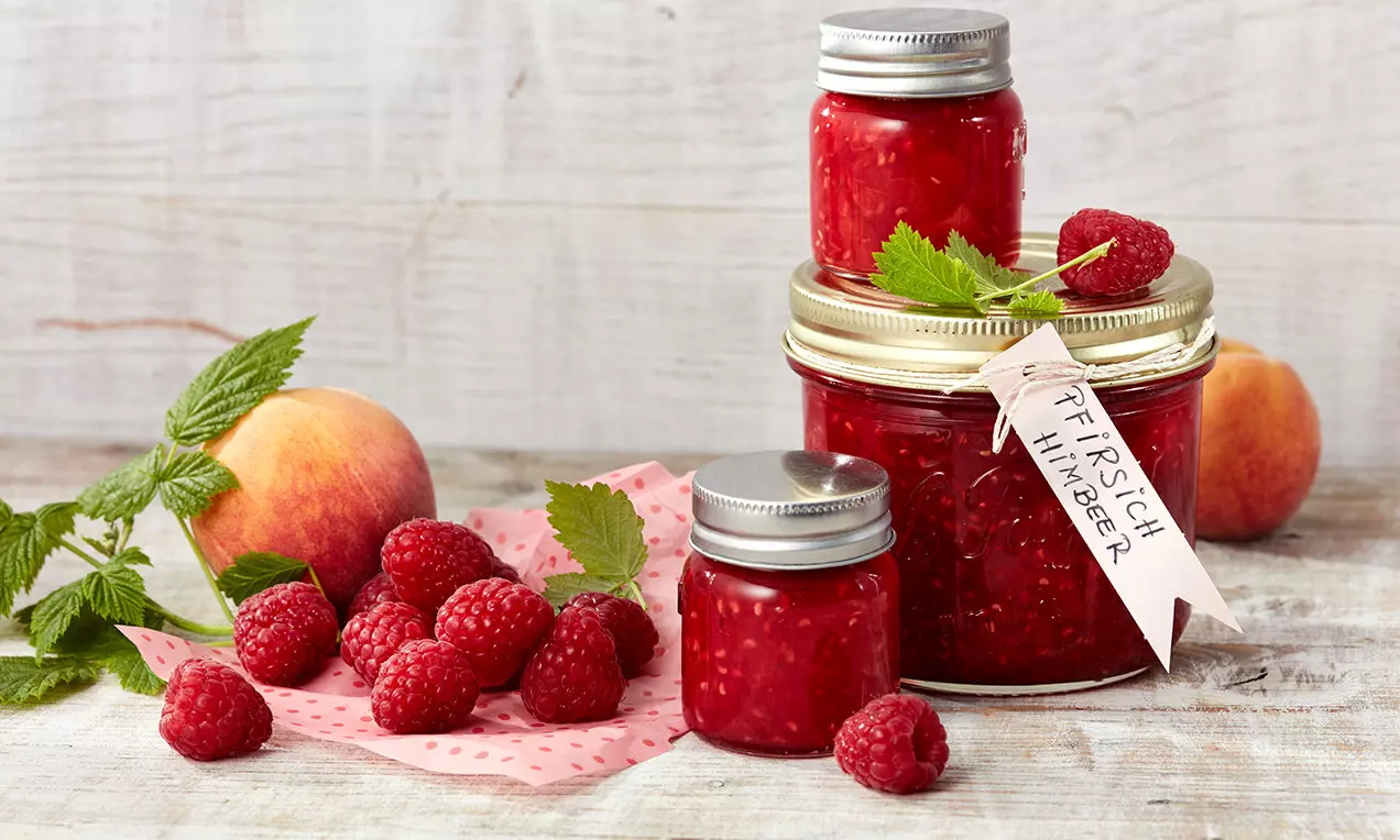 Буркани с наситено червено сладко със семенца от малини и праскови, украсени със зелени листа, заобиколени от свежи плодове върху дървена повърхност