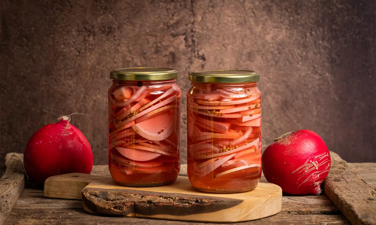 Червени и бели резени ряпа с пръстени лук в златиста течност, плътно подредени в прозрачни буркани върху дървена основа
